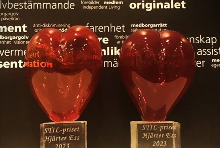 Två röda glashjärtan bredvid varandra. På den ena står STIL-priset Hjärter Ess 2023 Margareta Persson och på den andra STIL-priset Hjärter Ess 2023 Susanne Berg. En svart vägg med vita ord i bakgrunden.