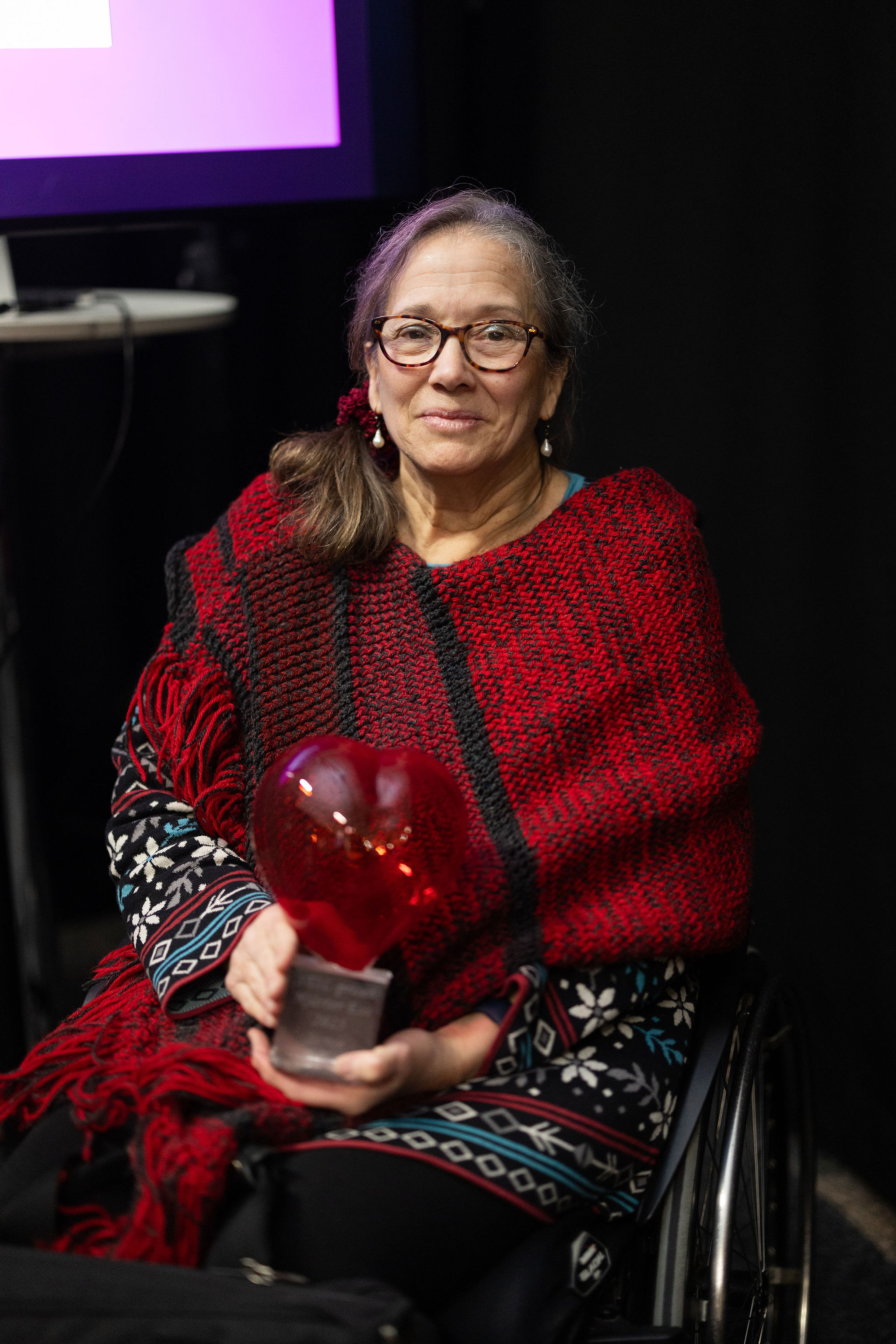 En kvinna i rullstol sitter på en scenliknande plats, iklädd en röd och svart poncho och håller i ett hjärtformat pris. Personen tittar mot kameran och ler.