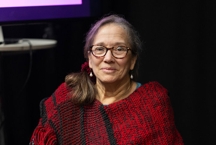 En kvinna i rullstol sitter på en scenliknande plats, iklädd en röd och svart poncho och håller i ett hjärtformat pris. Personen tittar mot kameran och ler.